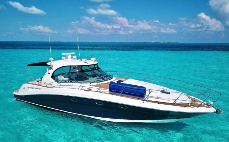 Port side of 44 ft Sea Ray 420 Sundancer luxury yacht charter anchored in stunning turquoise Caribbean, dark navy hull, teak decks, white superstructure, blue float on bow, Isla Mujeres in distance