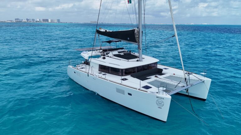 Vista aérea de estribor del catamarán Lagoon Beluga 45 ft fondeado en aguas turquesas del Caribe frente a Cancún, casco completo, mástil y trampolín blanco visibles, charter de lujo
