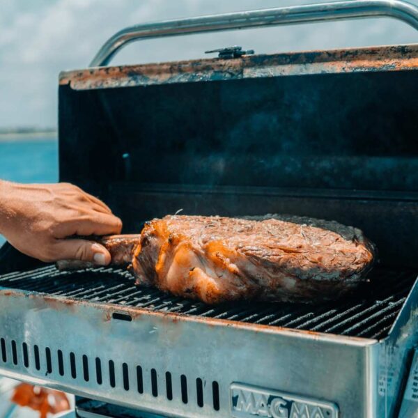 Hand turning a seasoned tomahawk steak on a stainless Magma grill mounted aboard a boat over the blue Caribbean.