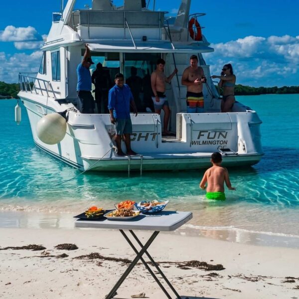 White table set with seafood platters on a Cancun beach beside an anchored yacht in clear turquoise shallows.