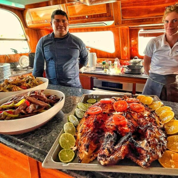 Smiling private chef and crew member presenting whole grilled fish with citrus and fajitas in the yacht galley.