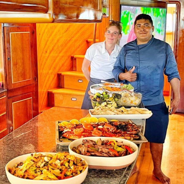 Cheerful chef and crew posing beside a tiered display of fajitas, grilled fish, pasta and salads aboard a yacht.