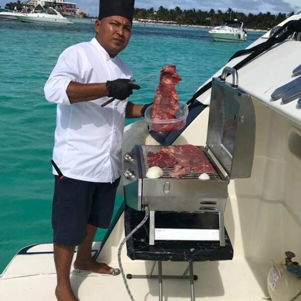Chef in white uniform grilling beef steaks on a boat deck with Cancun hotel zone and turquoise sea behind.