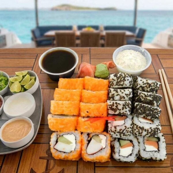 Assorted sushi rolls with soy sauce, wasabi and toppings served on a teak yacht table above turquoise water.