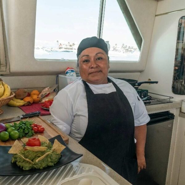 Smiling chef in the catamaran galley with a basket of fresh fruit and a plate of guacamole and nachos.