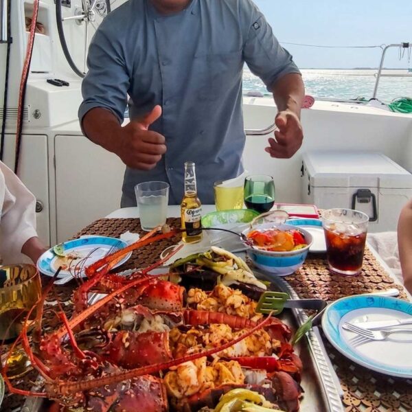 Beaming private chef giving a thumbs up behind a huge platter of grilled lobster, steak and vegetables on deck.