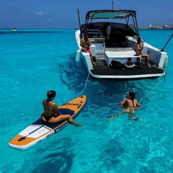 Guest rests on an inflatable paddleboard beside the swim platform while others snorkel off a yacht in bright turquoise water.