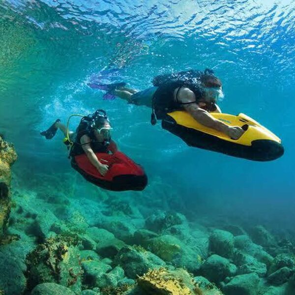 Two snorkelers ride underwater seabob scooters above a rocky reef, exploring colorful Caribbean marine life with mask and gear.