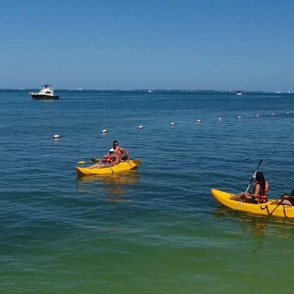 Two yellow tandem kayaks with guests in life jackets paddle the calm Cancun lagoon near an anchored boat.