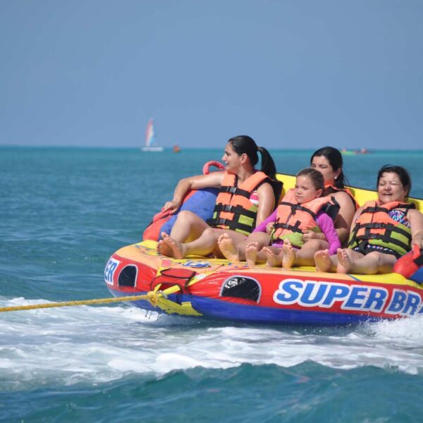 Family in life jackets rides the Super Brain inflatable towable tube, splashing across the Caribbean behind a Cancun boat.