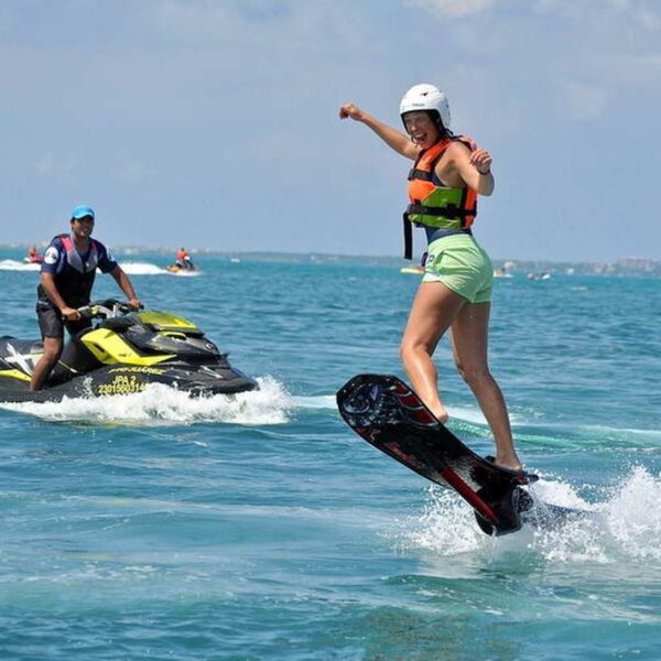 Smiling woman jetboards on a powered surfboard while an instructor follows on a jet ski in turquoise Cancun water.