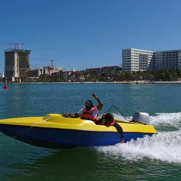 Two riders steer a small yellow speed boat across the Cancun lagoon with the hotel zone skyline behind them.
