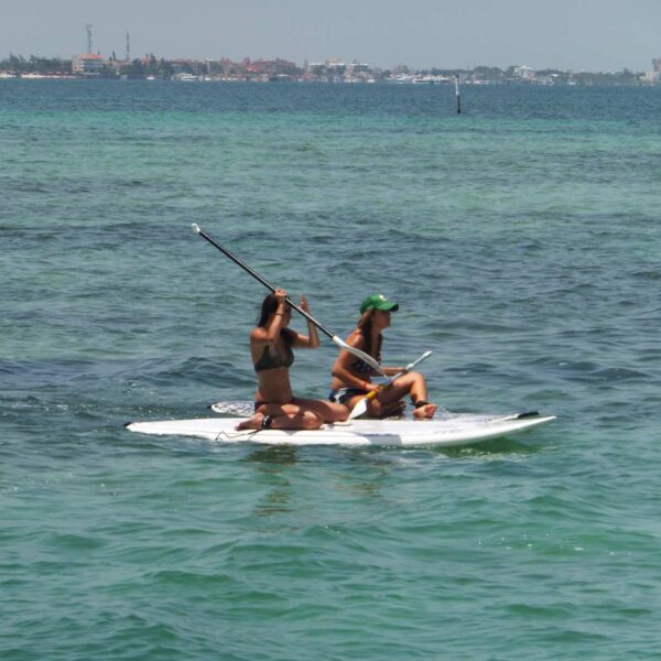 Two women share a stand up paddleboard on the calm Cancun lagoon with the city shoreline in the distance.