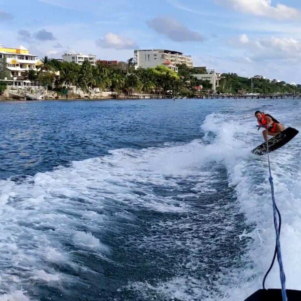 Wakeboarder in a life jacket carves through the boat wake along the Isla Mujeres shoreline on a sunny day.