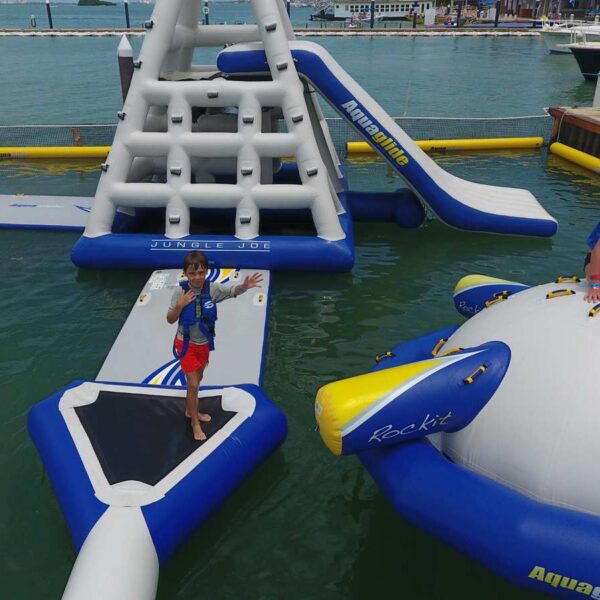 Children in life jackets play on a floating Aquaglide inflatable water park with a climbing tower, slide and bouncer in Cancun.